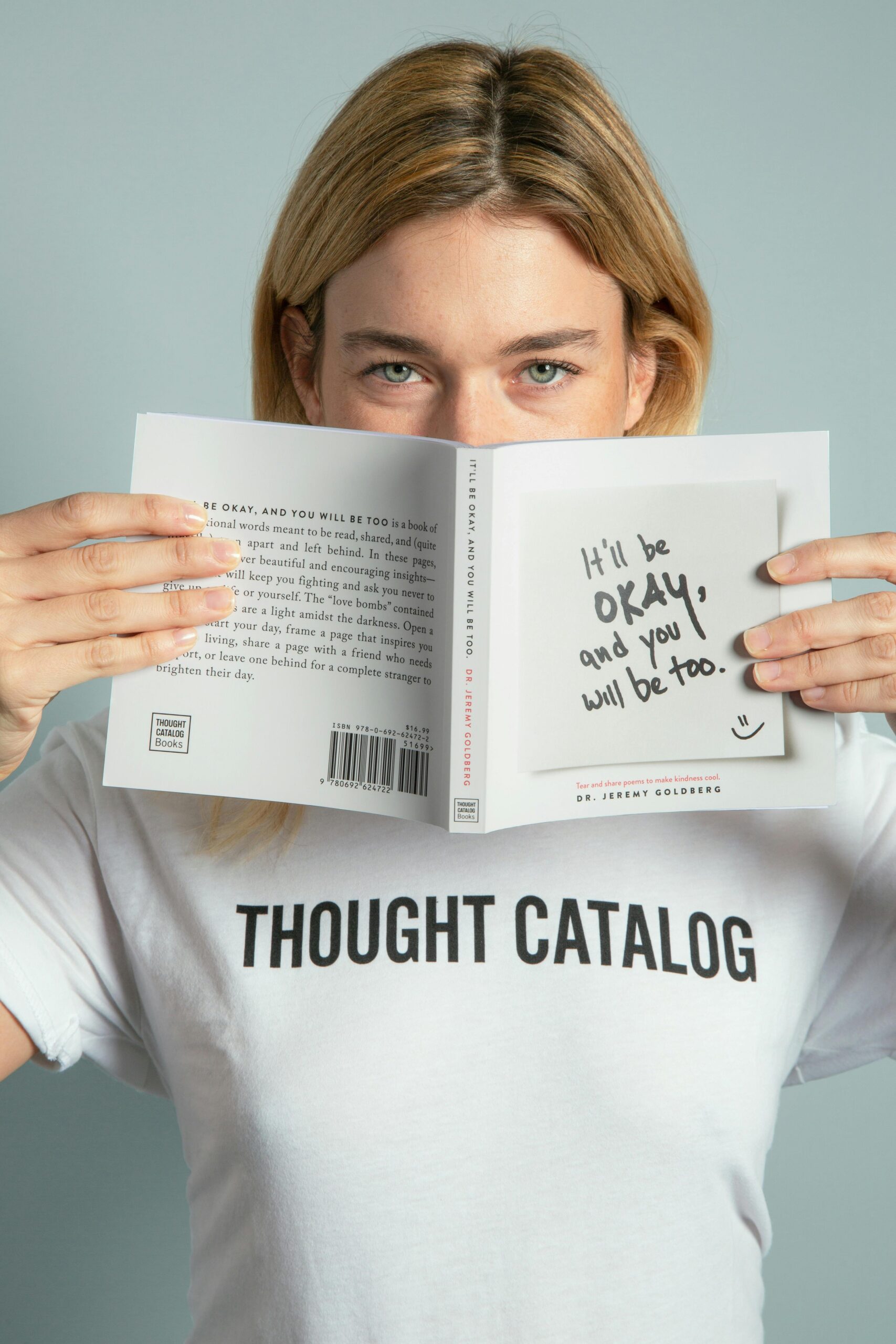 A person wearing a white "THOUGHT CATALOG" t-shirt holds a book titled "It'll Be OKAY, and You Will Be Too" by Dr. Jeremy Goldberg in front of their face, revealing only their eyes and forehead. A sticky note on the book cover repeats the title with a smiley face. The image conveys a message of reassurance and emotional support.