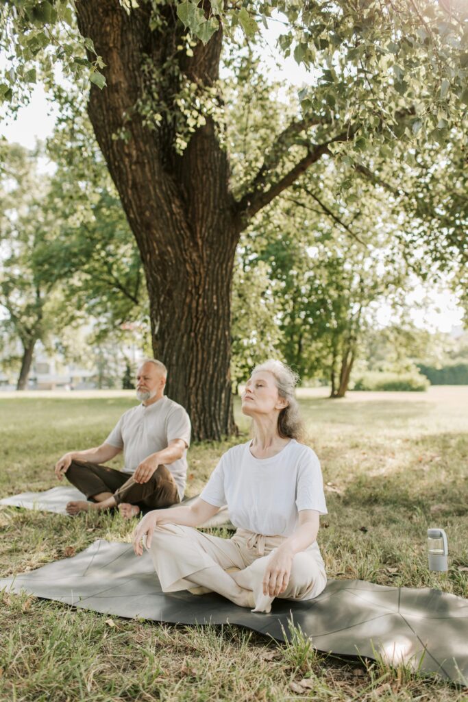 Two older adults practicing mindfulness meditation outdoors under a tree, seated cross-legged on yoga mats in a peaceful park setting.