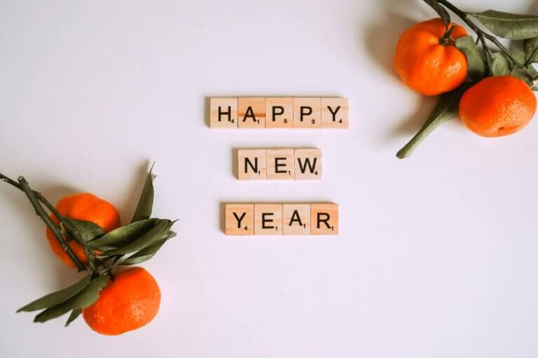 Festive New Year arrangement with 'HAPPY NEW YEAR' spelled in wooden Scrabble tiles, surrounded by fresh tangerines with green leaves on a clean white background.