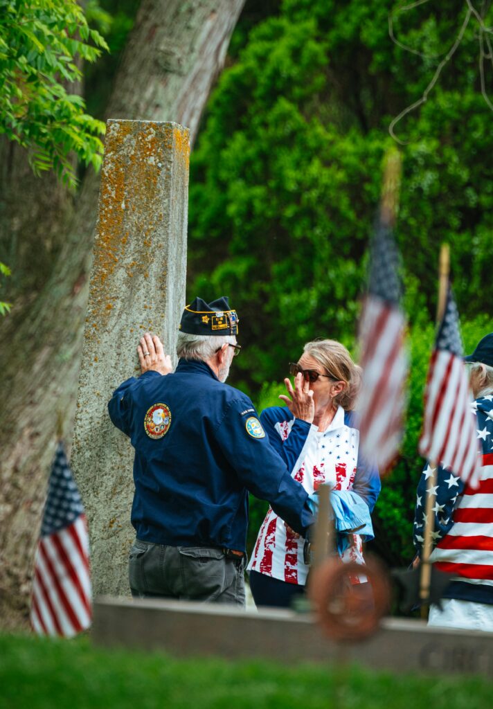 Veteran and woman sharing a heartfelt moment of remembrance near a monument on Memorial Day, surrounded by American flags and greenery.